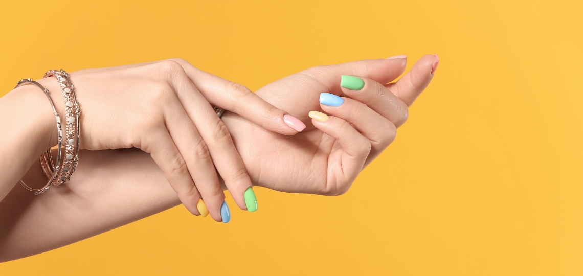 Eleganckie dłonie z kolorowymi paznokciami w pastelowych odcieniach na żółtym tle. Na zdjęciu widoczne są zadbane dłonie z manicurem w odcieniach mięty, błękitu, różu i żółci, ozdobione delikatnymi bransoletkami. Zdjęcie ma charakter ilustracyjny do artykułu o łyżeczkowatych paznokciach, jednak sama fotografia przedstawia zdrowe paznokcie i służy wyłącznie jako element graficzny tekstu. Profesjonalny manicure w wiosennych kolorach podkreśla elegancję kobiecych dłoni.