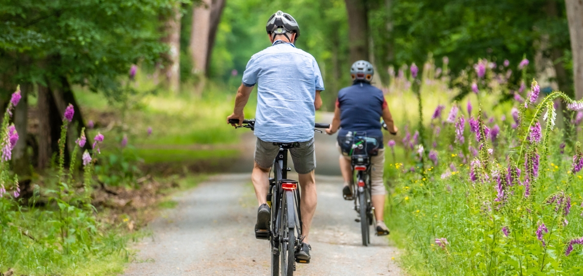 Dwie osoby jadące na rowerach leśną ścieżką otoczoną bujną, zieloną roślinnością i fioletowymi kwiatami. Zdjęcie ilustracyjne do podrozdziału o torbieli Bakera a jeździe na rowerze. Fotografia przedstawia aktywność fizyczną, która może być zarówno korzystna jak i problematyczna dla osób z torbielą podkolanową. Obraz ma wyłącznie charakter poglądowy i nie przedstawia pacjentów z tą jednostką chorobową, a jedynie wizualizuje formę aktywności omawianą w kontekście terapii i ograniczeń związanych z torbielą Bakera. Ukazuje rowerzystów w kaskach jadących po malowniczej trasie w otoczeniu przyrody.